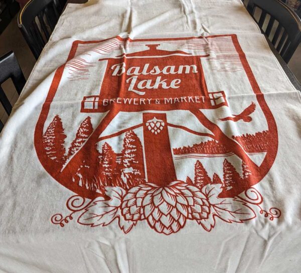 An White Beach Towel with the logo of balsam lake brewery & market, featuring hop cones and trees in a red and white color scheme.