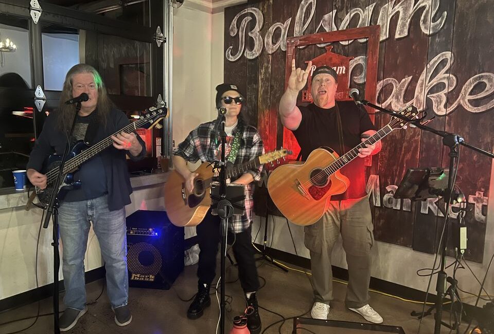 Three musicians performing on stage with acoustic guitars in front of a sign that reads "bojama baker.