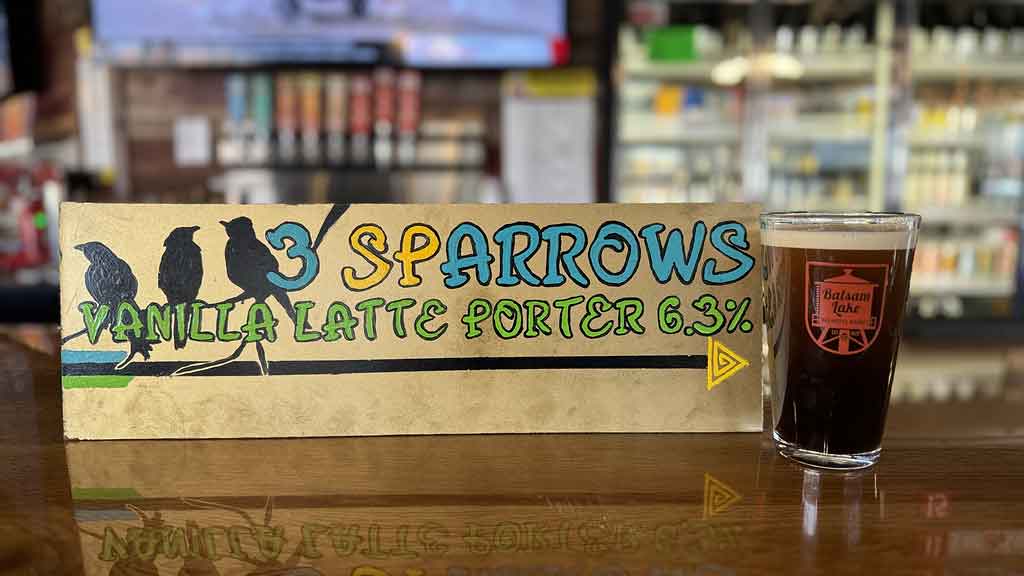 A glass of dark beer on a bar with a sign advertising "3 sparrows vanilla latte porter 6.3%" in the background.