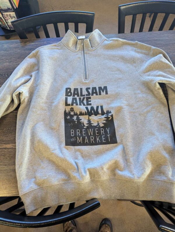 Quarter Zip Sweatshirt Grey displaying "Balsam Lake WI Brewery and Market" with a woodland graphic, situated on a wooden table.