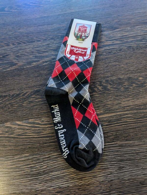 A pair of Brewery Socks in a black, red, and gray argyle pattern with "Scotland Forever" written on them, placed on a wooden surface.