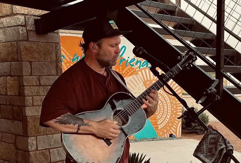 A person wearing a dark cap and maroon shirt plays an acoustic guitar while singing into a microphone. The background features a mural with vibrant patterns and text under a metal staircase.