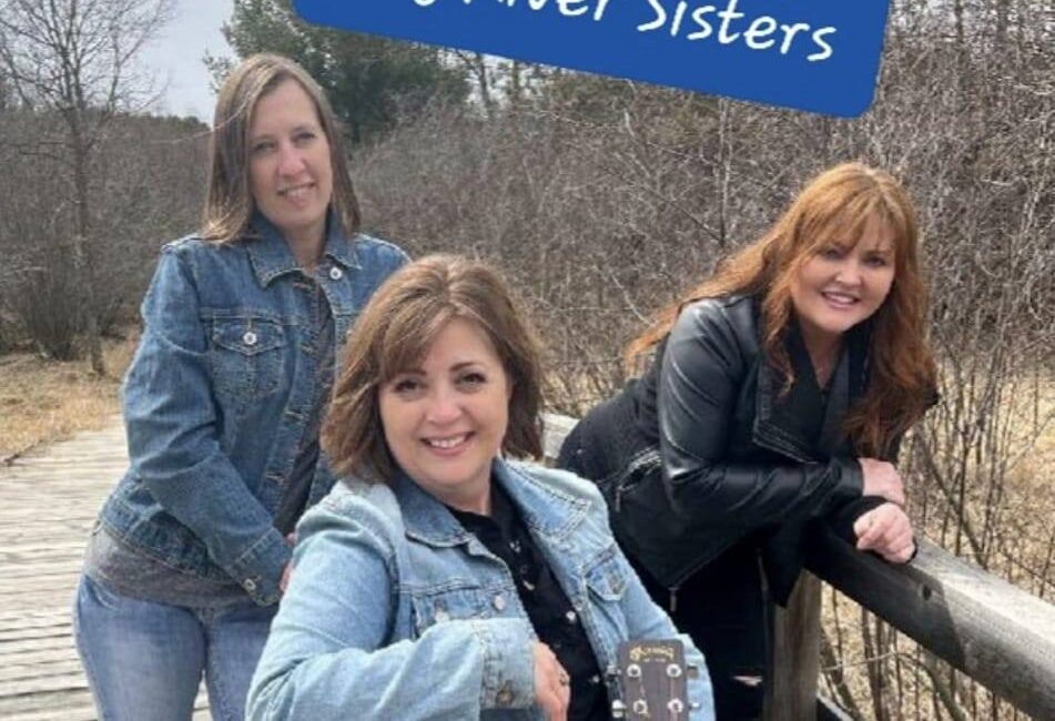 Three women, known as the Hay River Sisters, pose on a wooden boardwalk. One holds a guitar. Background includes trees and cloudy sky.