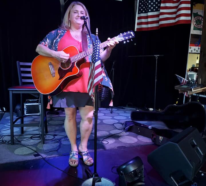 A woman stands on stage playing an acoustic guitar and singing into a microphone. An American flag is displayed on her mic stand and additional flags hang in the background. A tip bucket is by her feet.