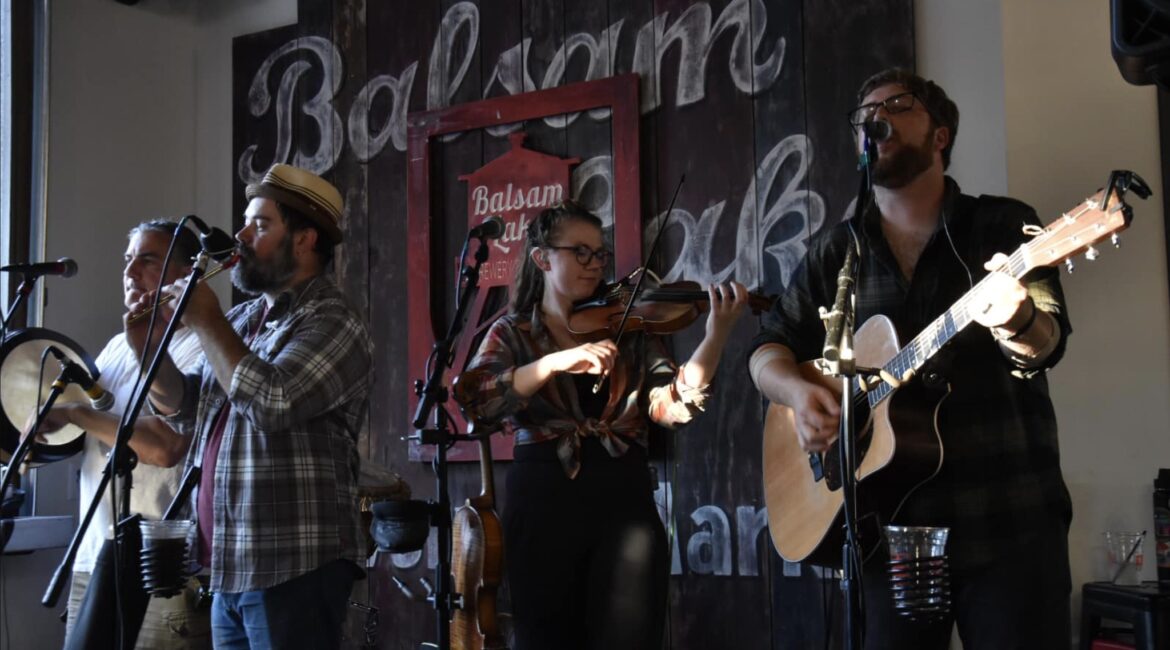 A band of four musicians performs with guitars, a violin, and a tambourine in front of a wooden wall with the name "Balsam Peak" displayed.