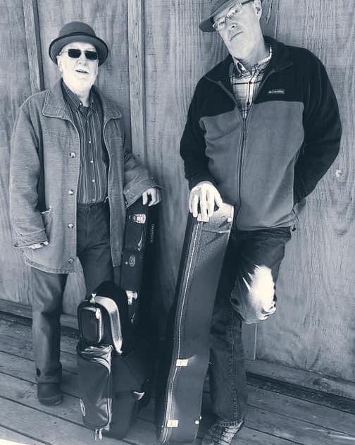 Two men standing against a wooden wall, each holding a guitar case, dressed in casual clothes and hats.