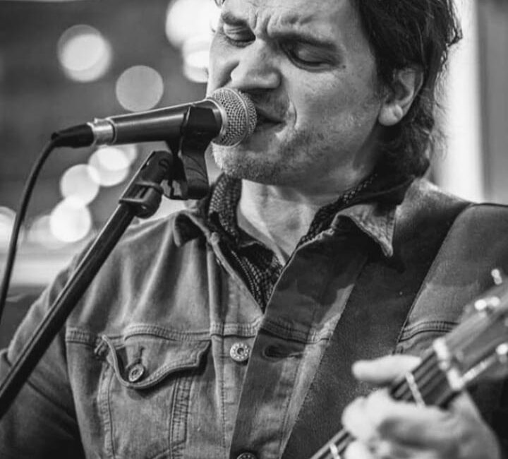 A man sings passionately into a microphone while playing an acoustic guitar. He is wearing a denim jacket. The background is blurred. Black and white photo.