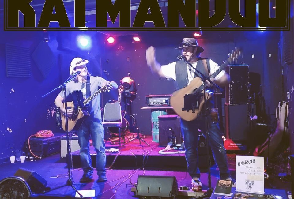 Two musicians with guitars perform on a stage under a "KATMANDUO" sign. They wear hats and casual attire, with various musical equipment around them. One musician gestures energetically.