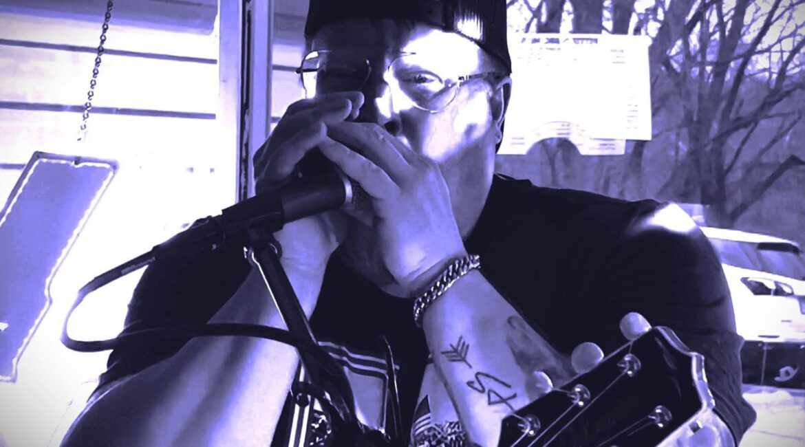 Person wearing a hat and glasses playing a harmonica into a microphone, with a guitar in the foreground.