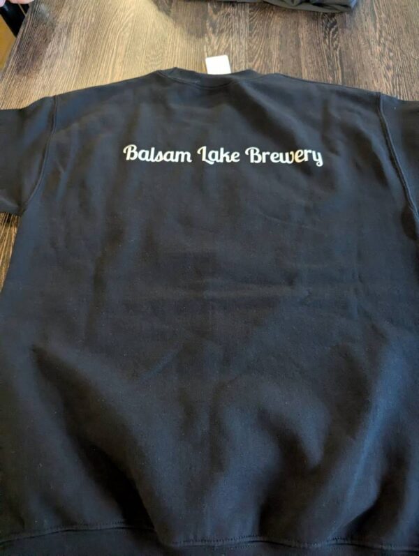 Black "Black Skull Hop Beard Tee" with "Balsam Lake Brewery" written in white on the back, lying on a wooden surface.