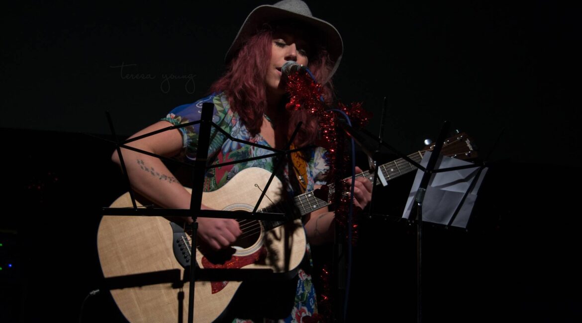 A musician with red hair plays an acoustic guitar and sings into a microphone on stage. She wears a floral dress and a grey hat. Music stands and decorations frame the scene.