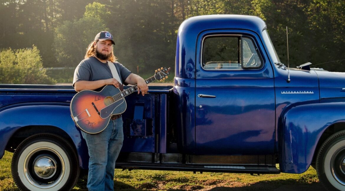 A man with a beard and long hair stands holding a guitar beside a vintage blue pickup truck in a grassy area with trees in the background.