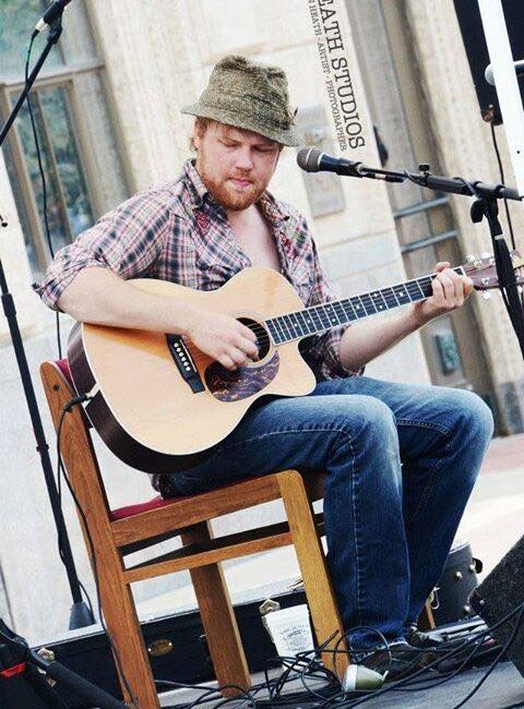 A man with a beard, wearing a hat and plaid shirt, sits on a wooden chair playing an acoustic guitar on an outdoor stage.