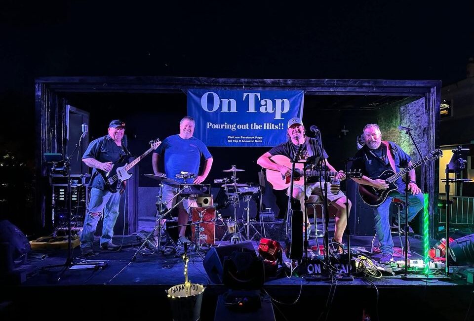 Four men are playing guitars and other instruments on an outdoor stage at night with a banner in the background that says "On Tap Pouring out the Hits!!".