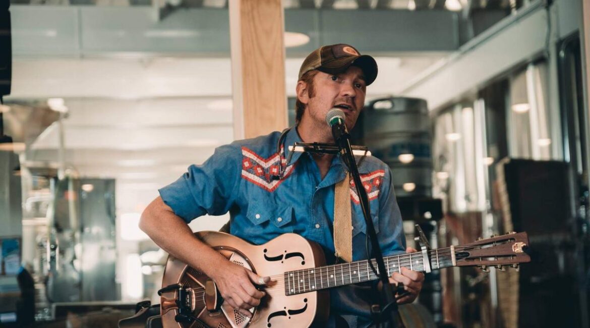 A musician in a blue shirt and cap plays guitar and harmonica while singing into a microphone in an indoor setting.