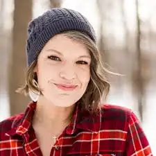 A person wearing a gray beanie and red plaid shirt smiles outdoors with trees in the background.