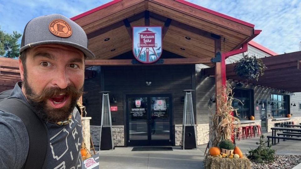 Man with a beard and mustache takes a selfie in front of a restaurant with a "Balsam Lake" sign. The building features wood accents, and there are pumpkins and plants near the entrance.