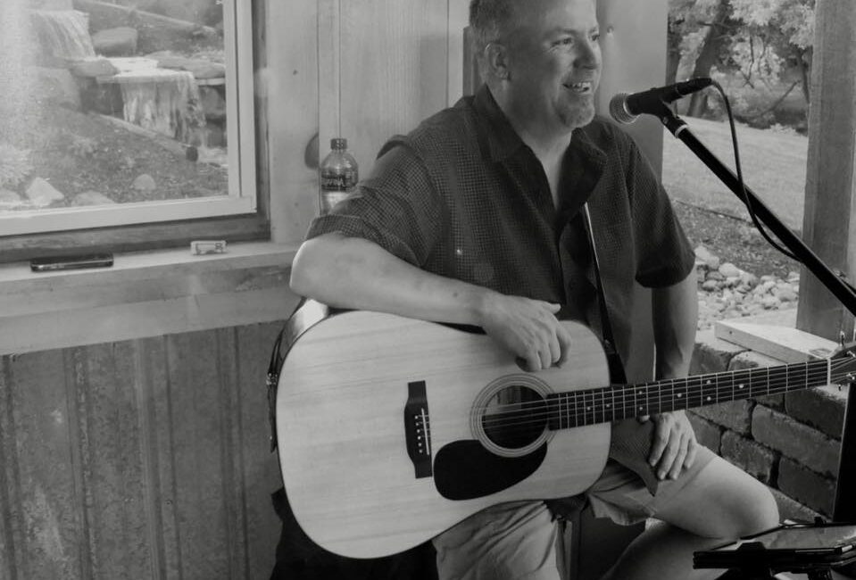 A man sits on a stool playing an acoustic guitar and singing into a microphone indoors, with a window showing an outdoor view.