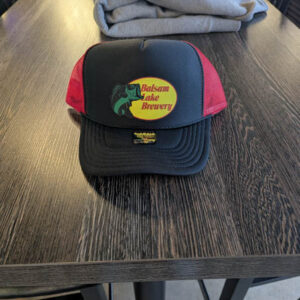 A red trucker hat featuring the "Balsam Lake Brewery" logo is placed on a wooden table.