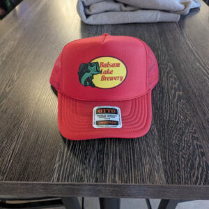 A Trucker Hat Bass Pro- Red featuring a "Balsam Lake Brewery" logo is resting on a wooden table, surrounded by folded clothes.