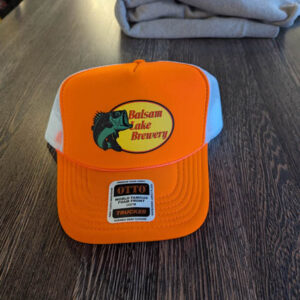 Red Trucker Hat from Bass Pro with Balsam Lake Brewery fish logo, placed on a wooden table in front of folded gray clothing.