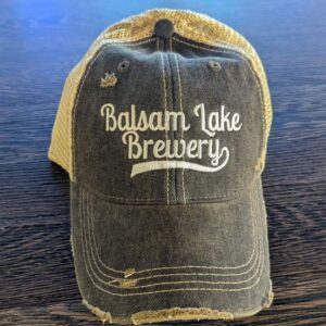 A "Distressed Baseball Cap Black" featuring "Balsam Lake Brewery" embroidered on the front, laid out on a wooden surface.