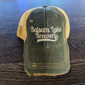 A Distressed Baseball Cap Green with "Balsam Lake Brewery" embroidered on the front is placed on a wooden surface.