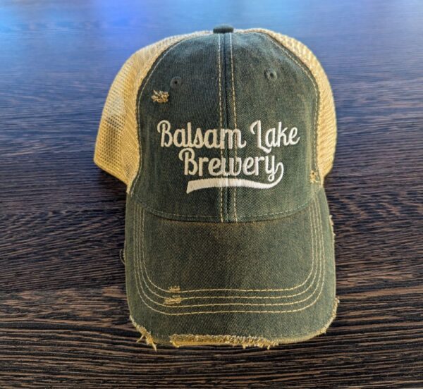 A Distressed Baseball Cap Green with "Balsam Lake Brewery" embroidered on the front is placed on a wooden surface.