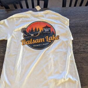 White tee on a table featuring a circular logo with "Balsam Lake Brewery & Market" and an eagle amidst trees and a lake.