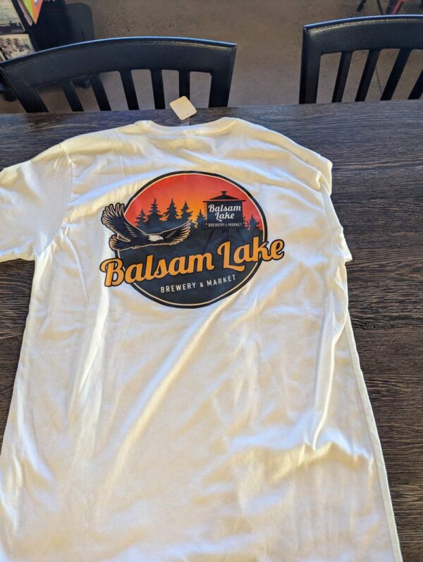 White tee on a table featuring a circular logo with "Balsam Lake Brewery & Market" and an eagle amidst trees and a lake.